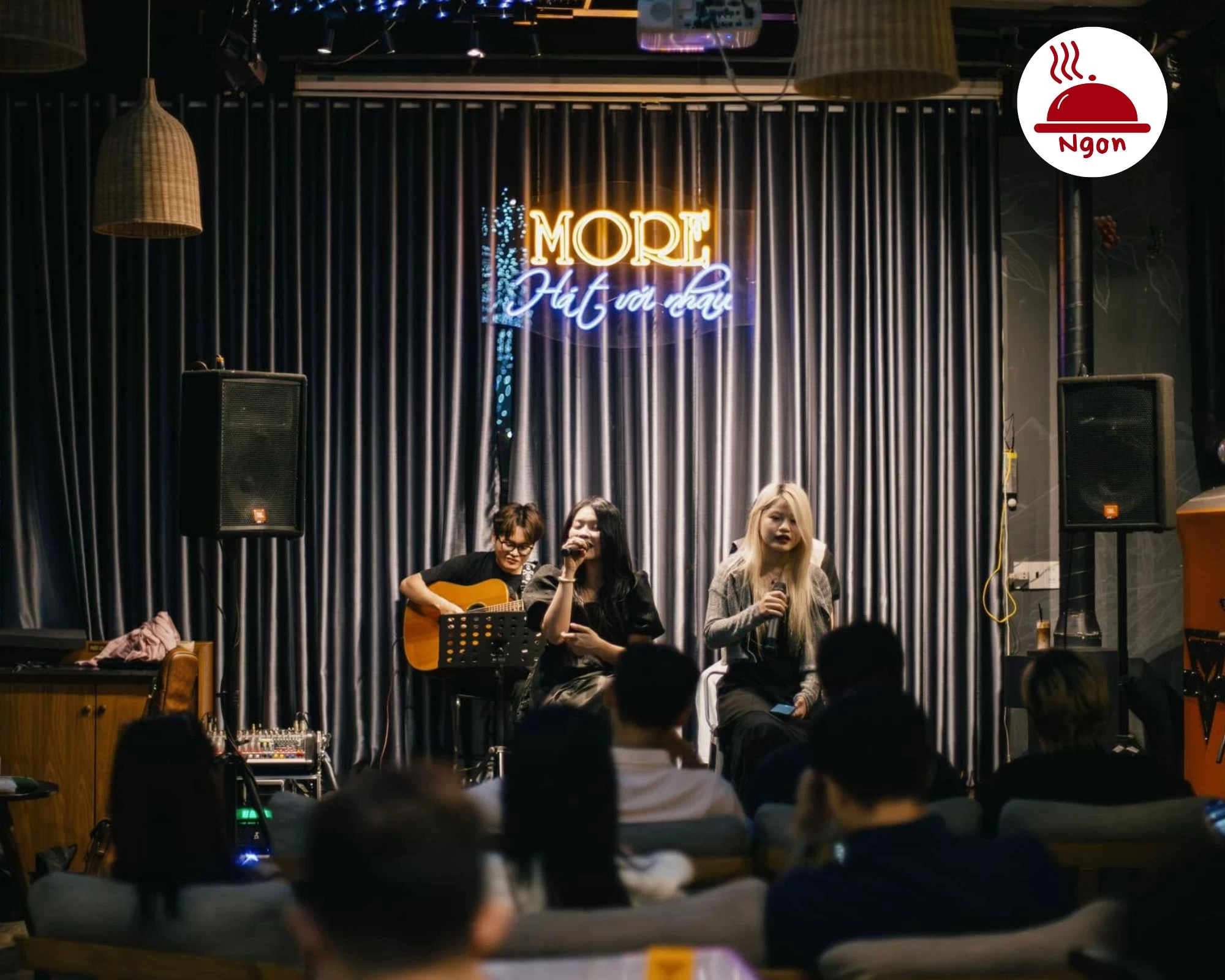 Trải nghiệm cà phê acoustic_ ban nhạc chơi live trên sân khấu nhỏ, đèn sân khấu và khán giả ngồi gần — không khí âm nhạc đặc trưng